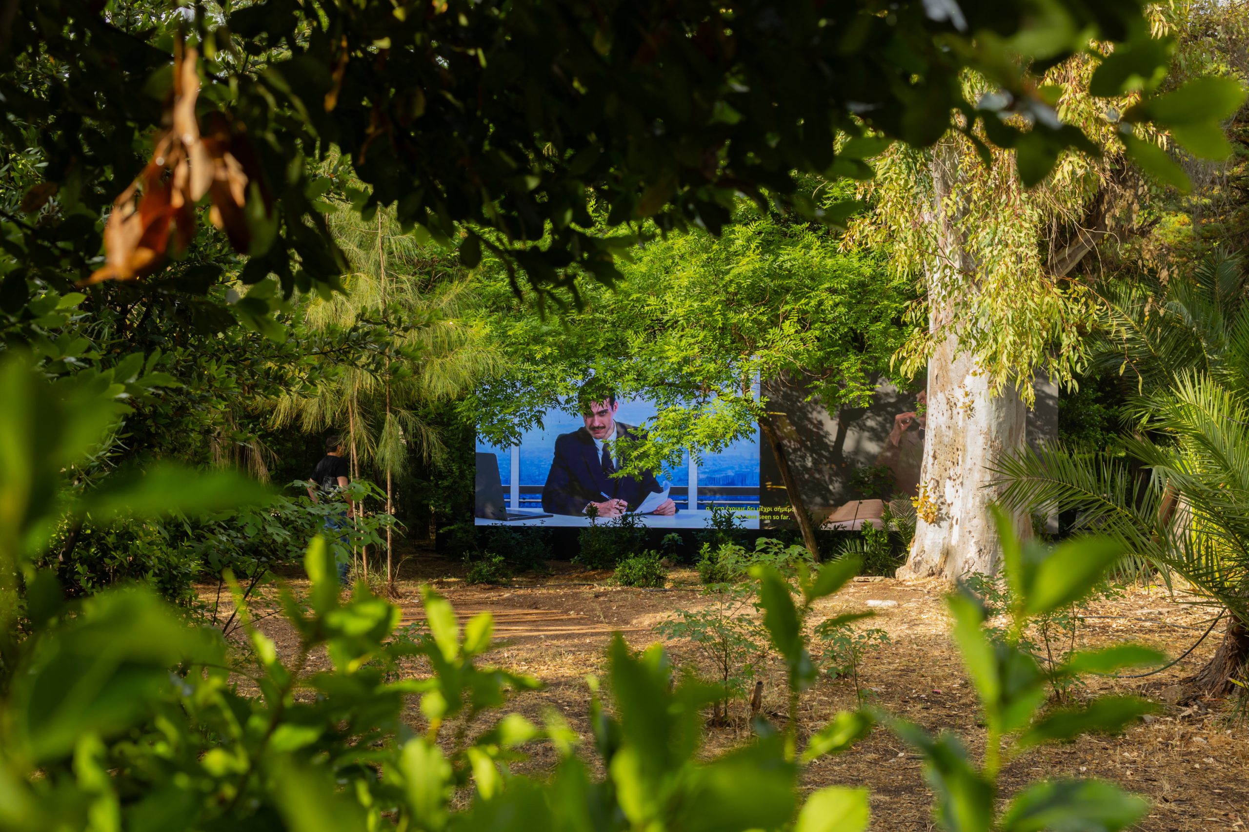 Aias Kokkalis - Ares Awakening - A TV displaying a man in a suit speaking is set outdoors, partly obscured by lush green trees and plants, blending into the garden like a CGI scene crafted in Houdini. Sunlight filters through the leaves.