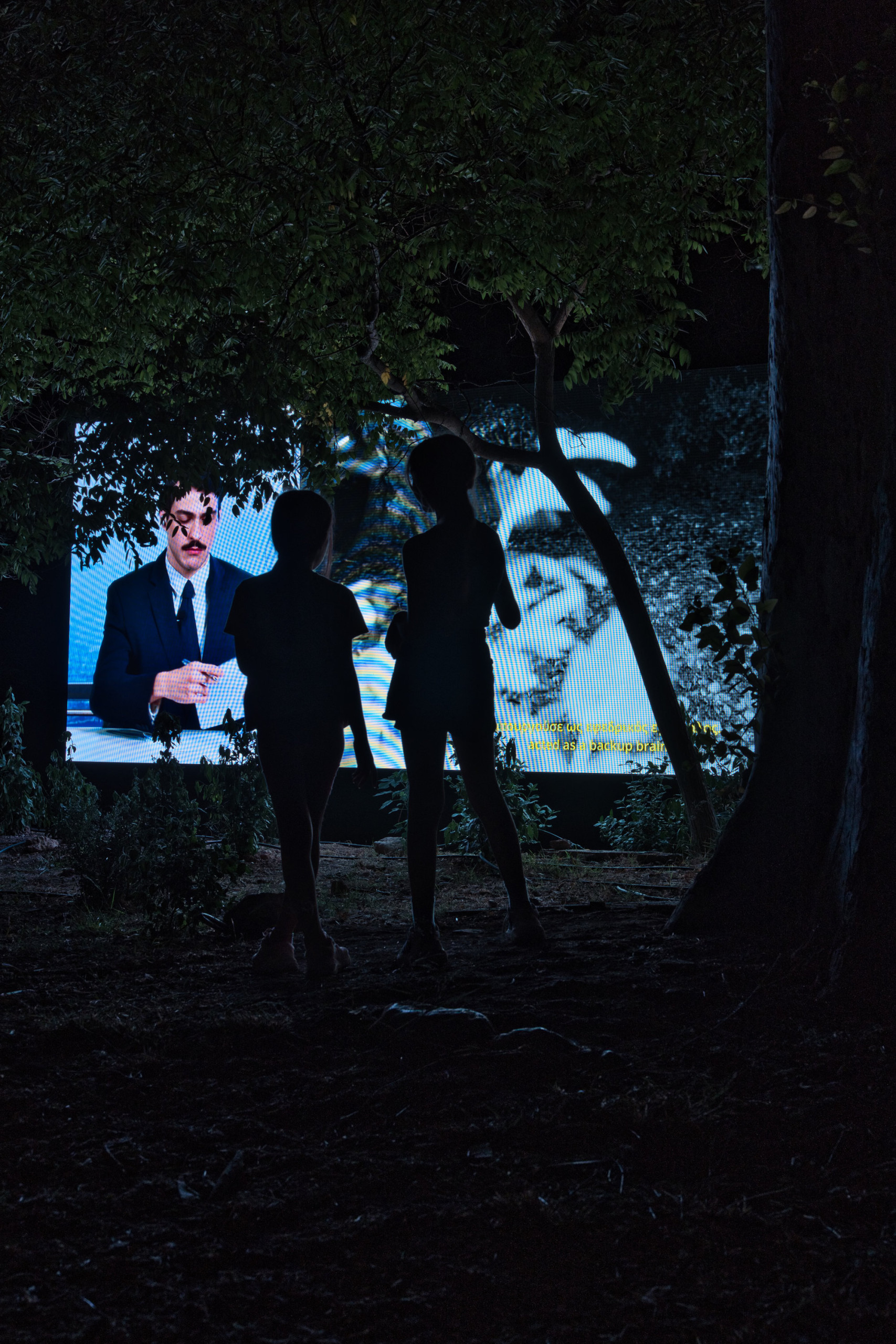 Aias Kokkalis - Ares Awakening - A group of people, including adults and children, stand and kneel outdoors at night, illuminated by bright lights among trees in a scene reminiscent of CGI Houdini artistry. Some look ahead with interest, while the background is dark and partly obscured by foliage.