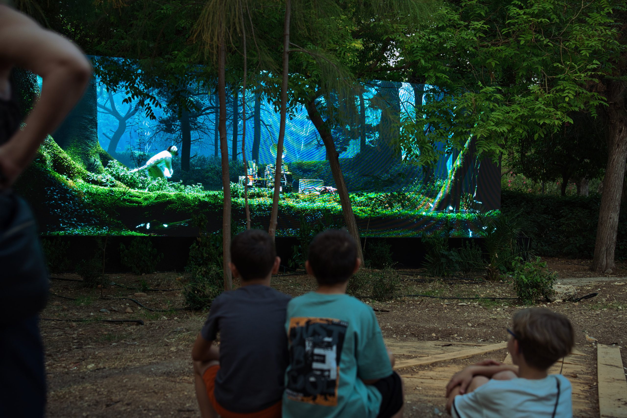 Aias Kokkalis - Ares Awakening - Three children sit outdoors on the ground, captivated by a large, bright digital screen displaying a vibrant forest scene with a mythical CGI creature and lush greenery. Tall trees and foliage surround this enchanting Houdini-powered viewing area.