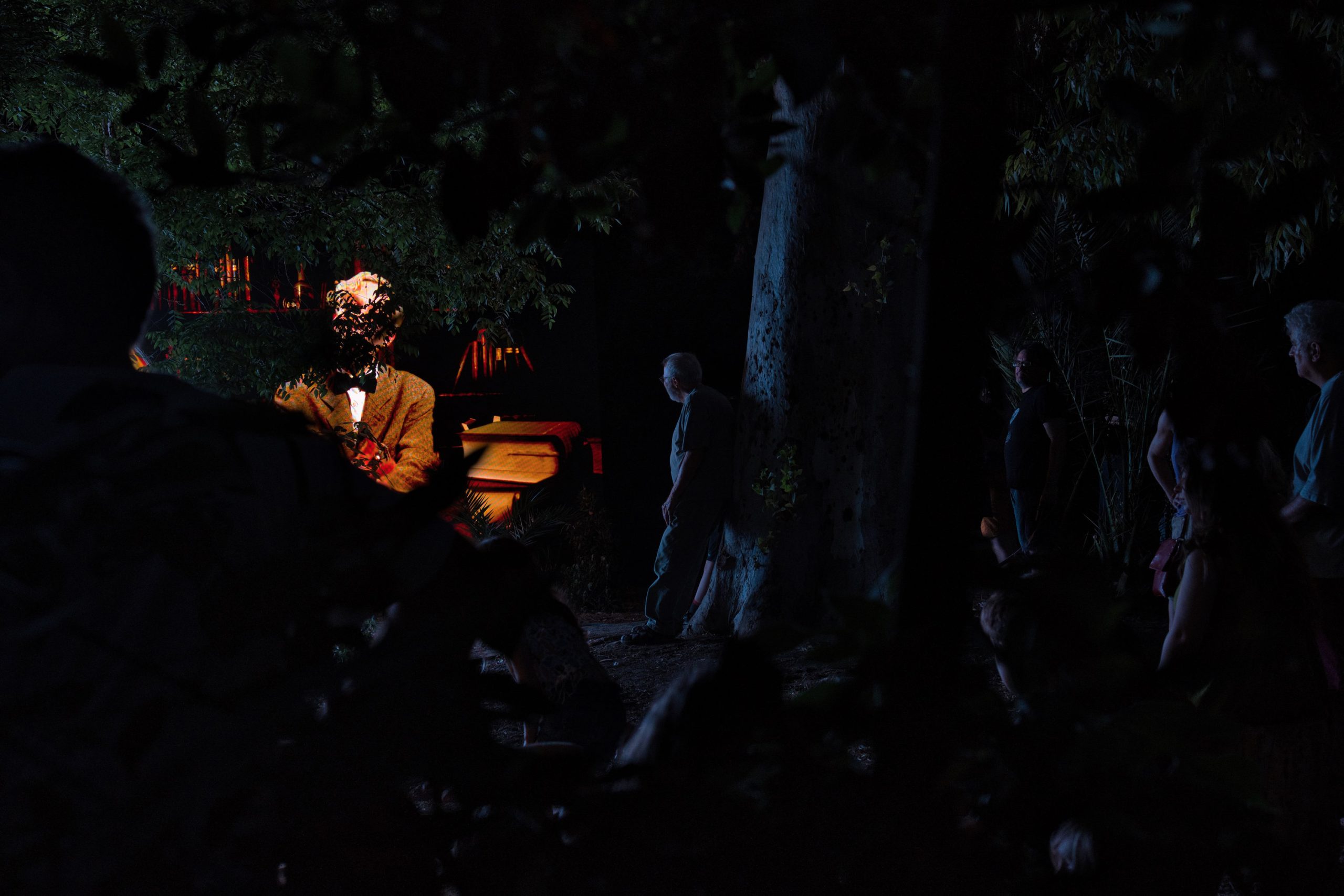 Aias Kokkalis - Ares Awakening - A dimly lit, posthuman scene at night with several people standing around. The focus is on a person with a bouquet, their face obscured by bright light, with trees and a parked car in the background. Shadows and darkness dominate the image.