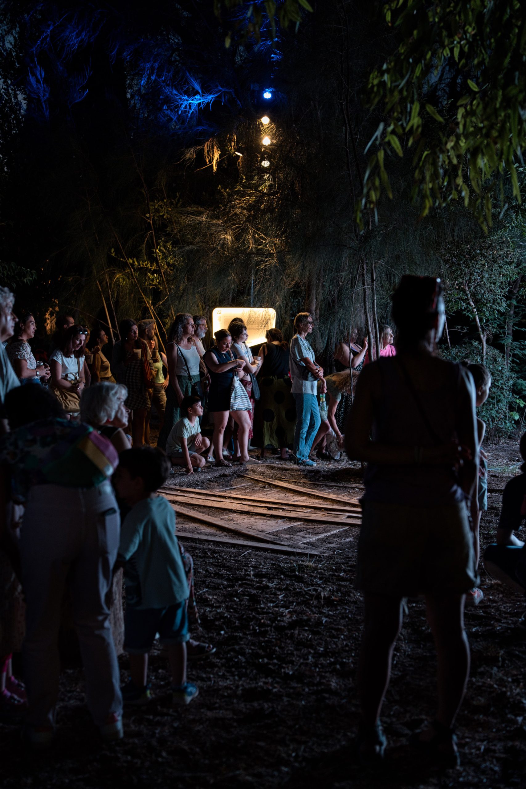 Aias Kokkalis - Ares Awakening - A group of people, including adults and children, stand and kneel outdoors at night, illuminated by bright lights among trees in a scene reminiscent of CGI Houdini artistry. Some look ahead with interest, while the background is dark and partly obscured by foliage.
